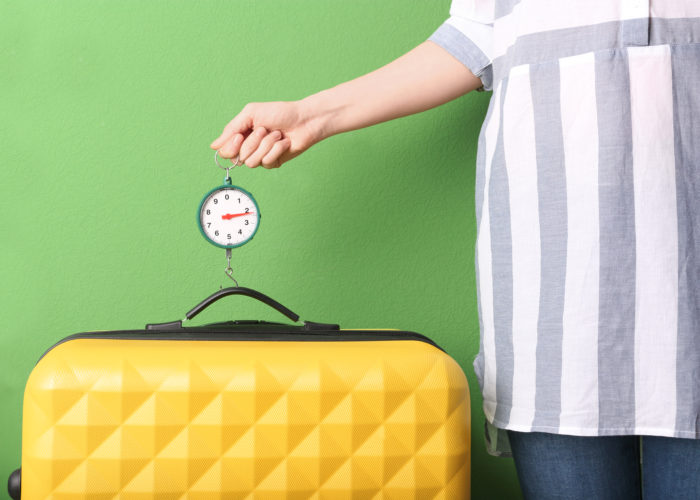 Close up of person wearing a dress and using a luggage scale to weigh a yellow suitcase against a bright green wall