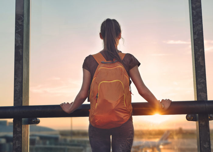 Woman standing with backpack looking out a window at sunset in an airprot