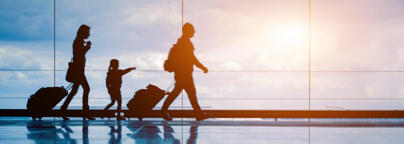 Silhouette of family walking along a glass-walled walkway in airport, rolling their suitcases