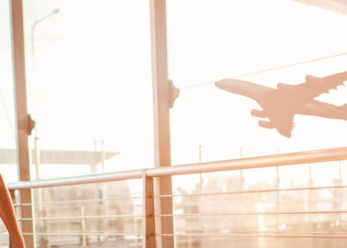 Woman pulling suitcase down hallway in airport next to glass window, out of which is a view of a plane taking off