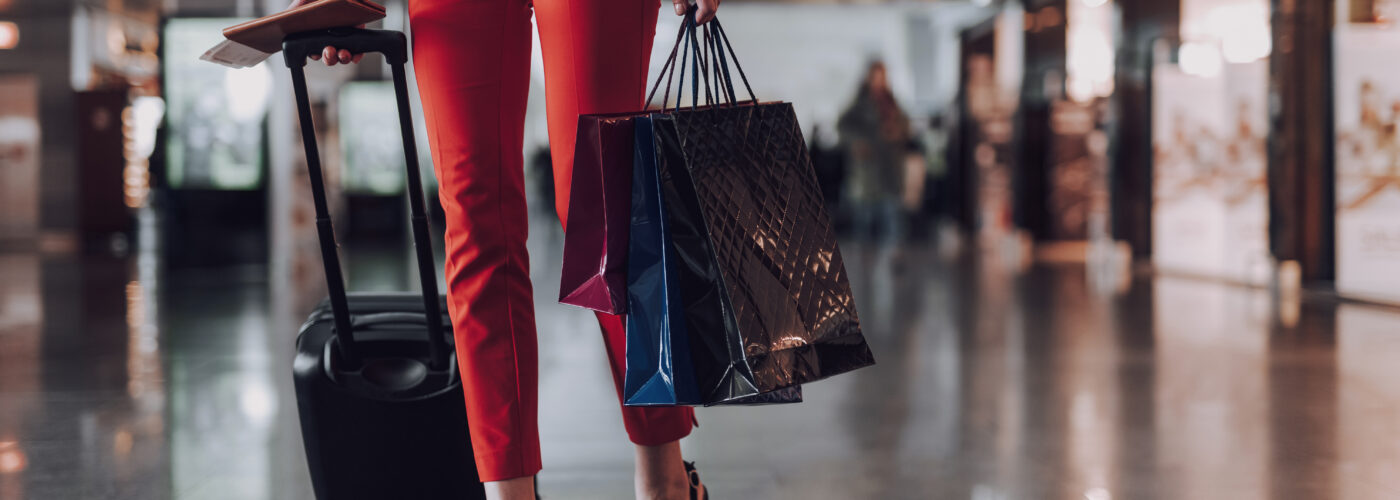 Woman, seen from the waist down, wearing high heels and carrying a rolling suitcase and shopping bags in an airport