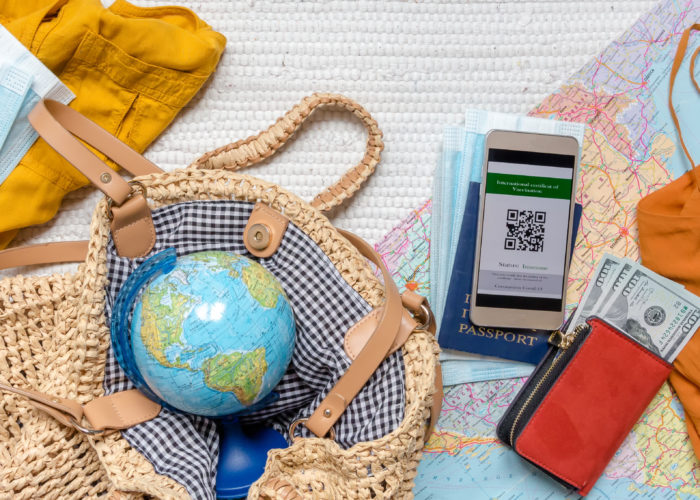 A travel themed flat lay featuring a small globe, face masks, a COVID-19 immunity passport, sunglasses, cash, and a passport